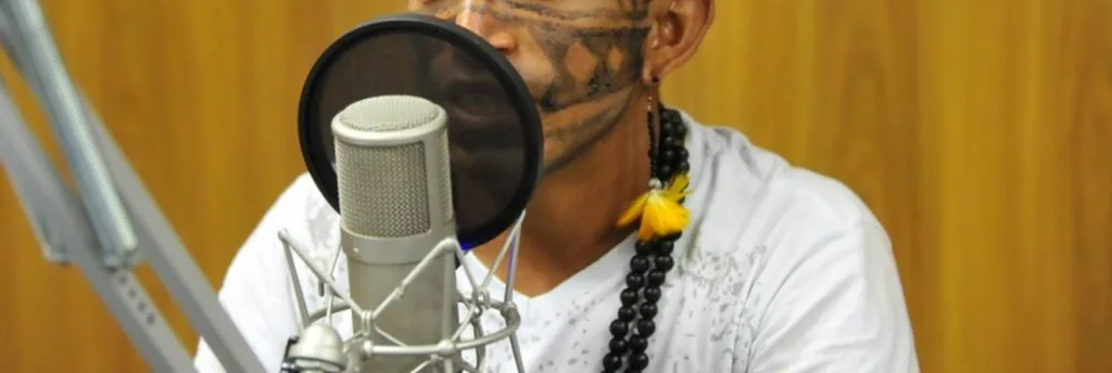 Jovem indígena, Valdenir Munduruku, com pinturas etnícas, cordão e camisa branca, falando em microfone de rádio.