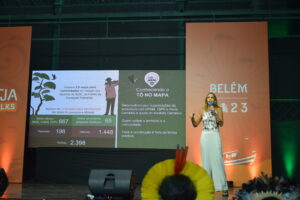 Isabel castro, mulher vestindo calça branca e blusa estampada, apresenta iniciativa Tô no Mapa em palco do 5ª PROTEJA Talks.