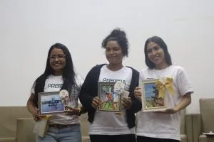 Três mulheres olham para câmera e sorriem enquanto seguram fotos premiadas.