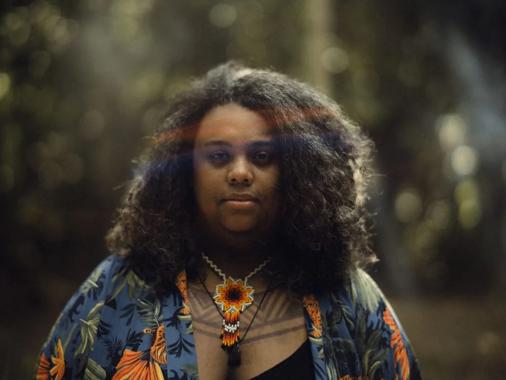 Retrato de Kamila Camilo. Uma mulher preta trajando uma camisa azul com detalhes de flores com pétalas laranjas. Ela usa um colar indígena no pescoço e um raio do sol repousa em sua cabeça. 