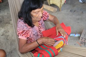 Anciã Imakor Paiter em oficina de confecção da tipoia, instrumento de algodão feito para carregar bebês. A anciã é uma senhora sentada em uma rede, manuseando a tipoia de algodão vermelho. Museu Paiter a Soe: resgatando a cultura para proteger a floresta 