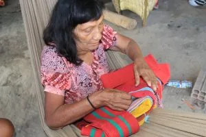 Anciã Imakor Paiter em oficina de confecção da tipoia, instrumento de algodão feito para carregar bebês. A anciã é uma senhora sentada em uma rede, manuseando a tipoia de algodão vermelho. Museu Paiter a Soe: resgatando a cultura para proteger a floresta 